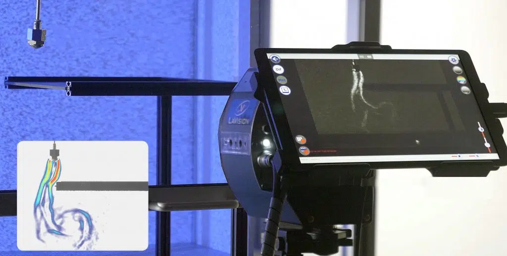 Close-up of a LaVision FlowMaster imaging system, showing a nozzle injecting fluid and a tablet displaying a Particle Image Velocimetry (PIV) visualization of the flow structures.